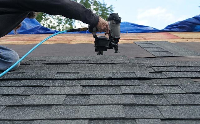 Worker using nail gun to install asphalt shingles on roof